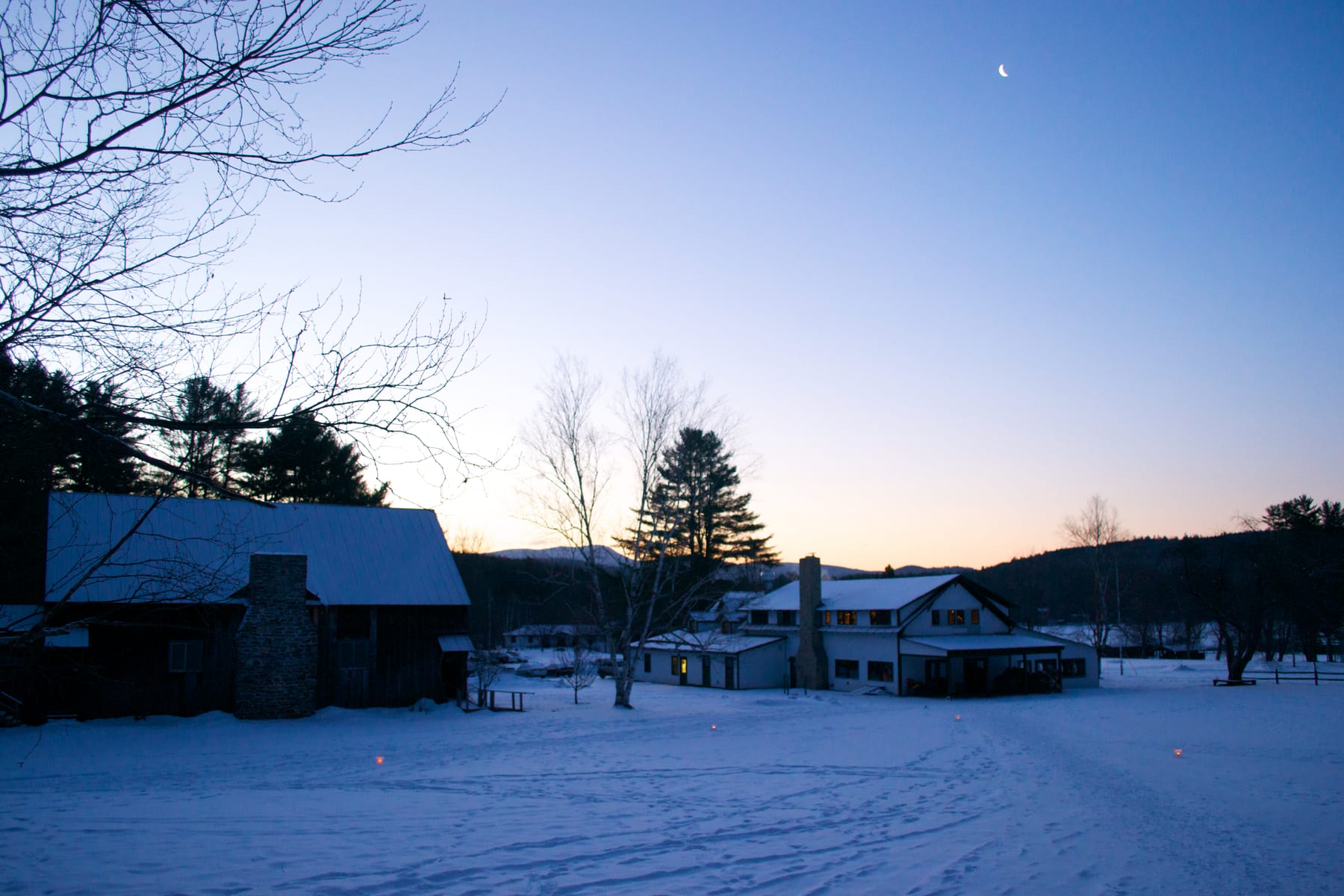 Snowy landscape with buildings and a moonlit sky at dusk.